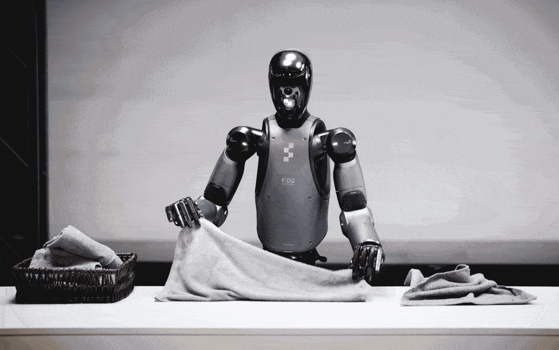 A silver and black bipedal humanoid uses its five fingered hands to gray towels. A towel to be folded sits on one side, and on the other is a basket holding folding towels.