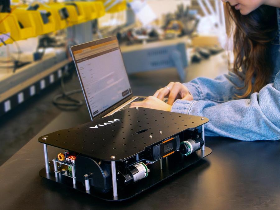 A woman sits at a laptop programming. In front of her is a robot consisting of two tiers of black rectangles with cameras and electronics in the middle.