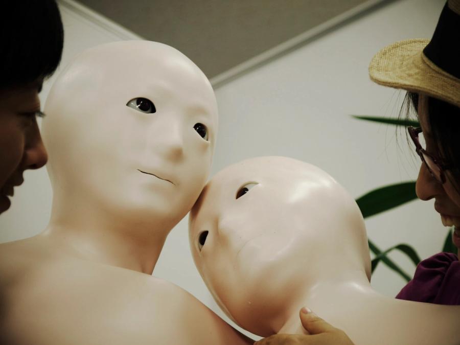 Two Telenoid robots with soft flesh-colored torsos and doll-like faces are pressed together.