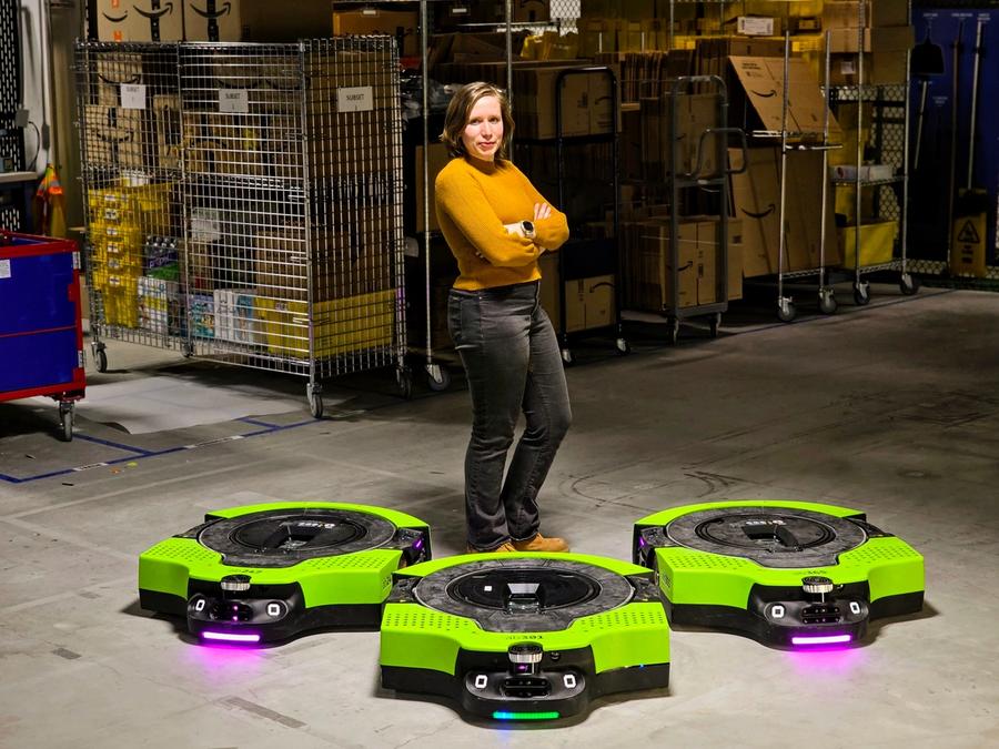 A blonde woman in a yellow shirt and black jeans stands in a warehouse. Three wide, low to the ground green and black Amazon warehouse robots are in front of her.