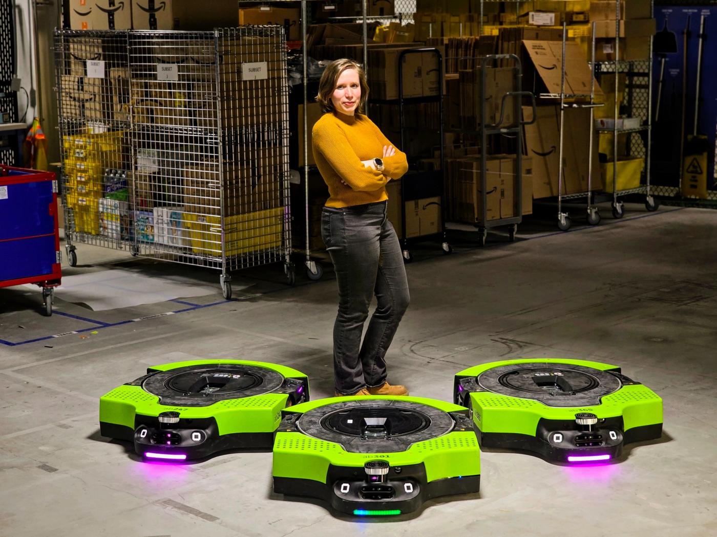 A blonde woman in a yellow shirt and black jeans stands in a warehouse. Three wide, low to the ground green and black Amazon warehouse robots are in front of her.