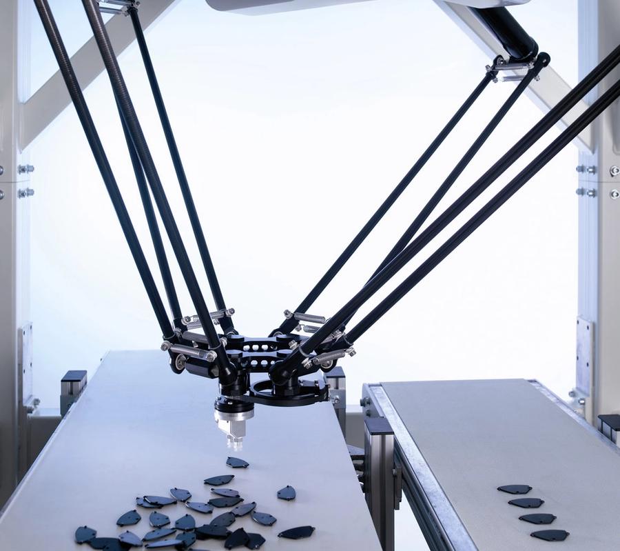 The robots four-arm rotational platform sits over a white conveyer belt with small black pieces.