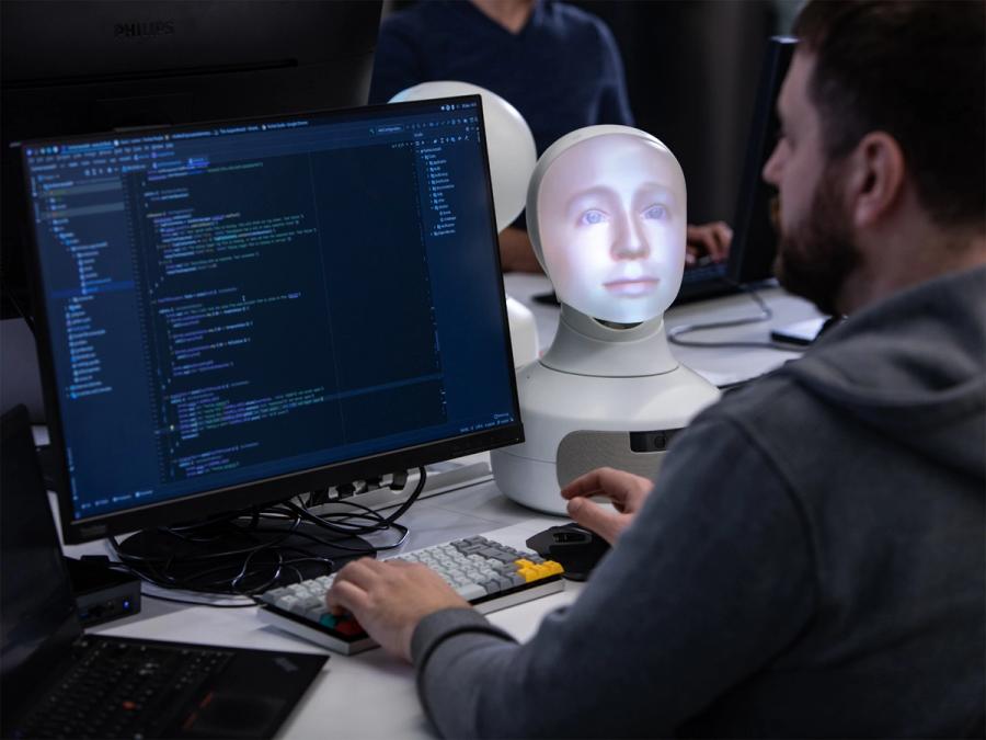 A man sits programming at a computer sitting next to a tabletop Furhat robotic head displaying a male face.