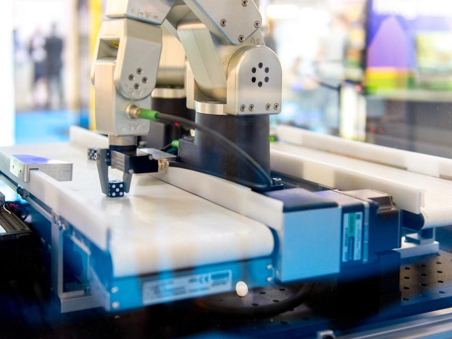 The robotic arm sits on a work tabletop and is bent over to grip a small die between its two fingers.