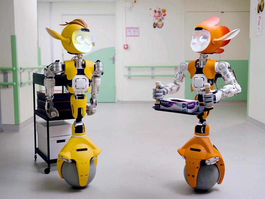 Two rolling humanoid robots with fox-like faces carry items in a hospital.