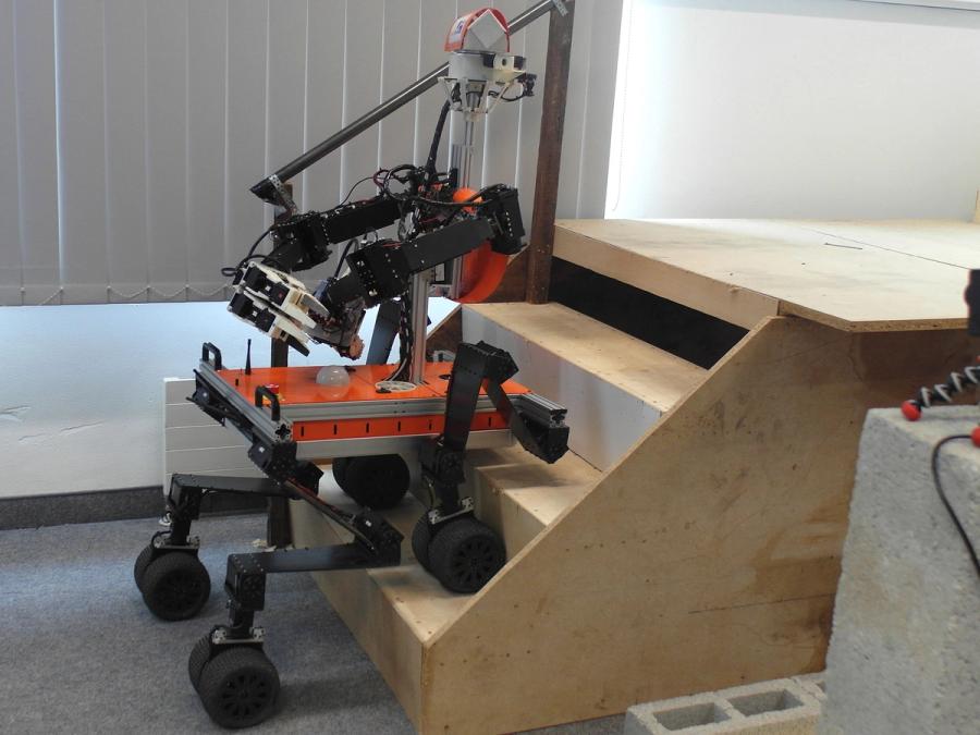 The orange and black robots back wheels are on the ground while its front legs are positioned on a stair.