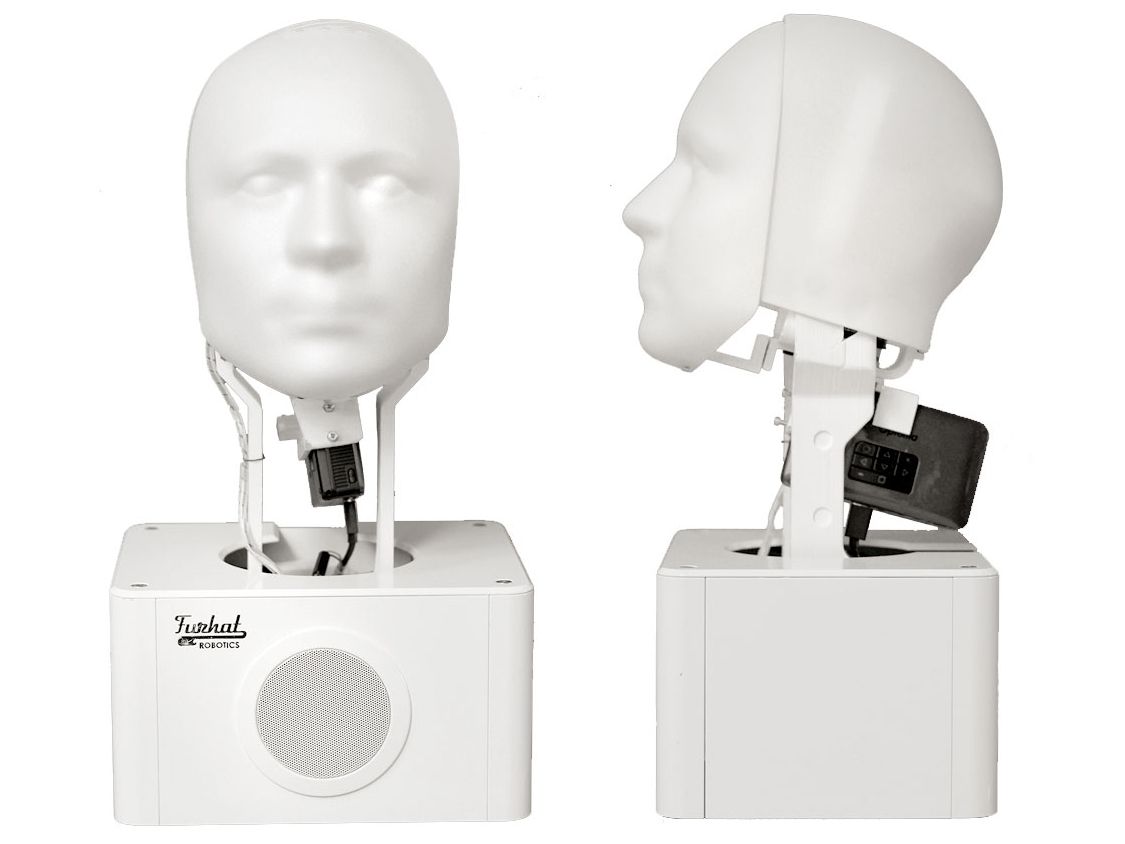The original Furhat robot head is seen from front and side view. It looks like a white box with a head shape held above it with some exposed electronics.