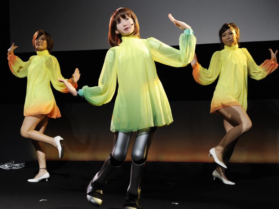 The female robot is seen in a dress dancing on stage and singing into a small microphone headset. Two human females are posed behind it, wearing matching outfits.