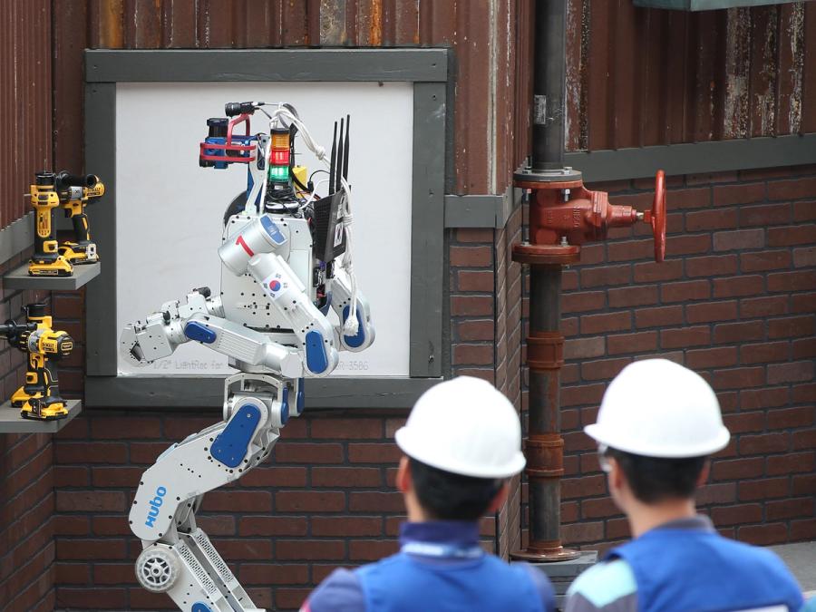 A bipedal humanoid robot stands in front of a shelf of drills while two people in hard hats look on.