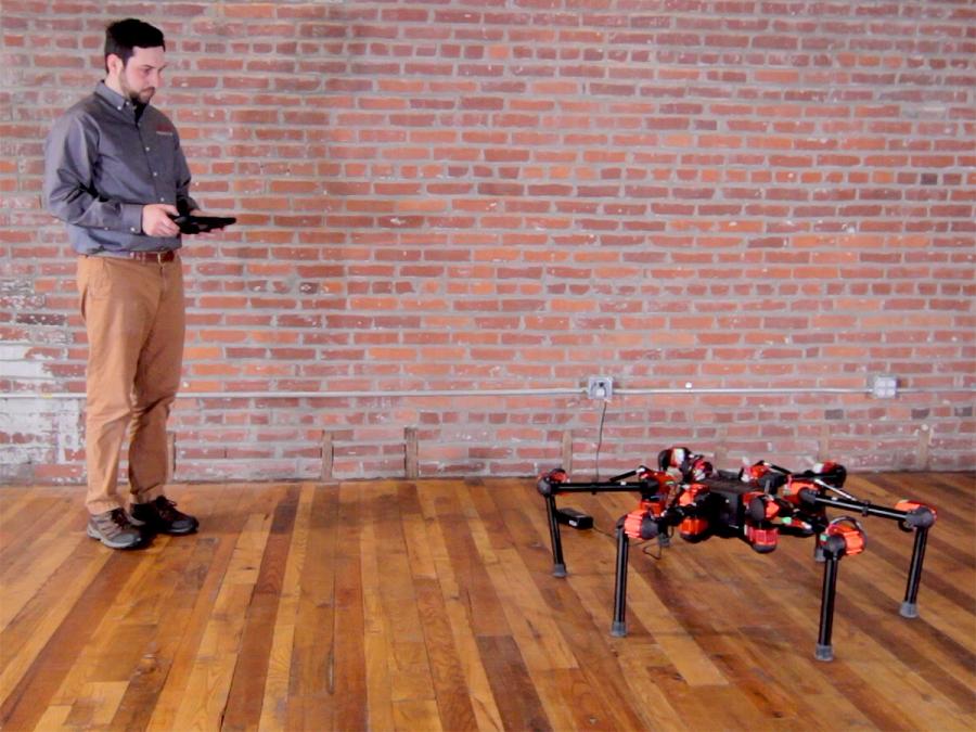 A man with a device stands looking at a red, black and silver hexapod robot.