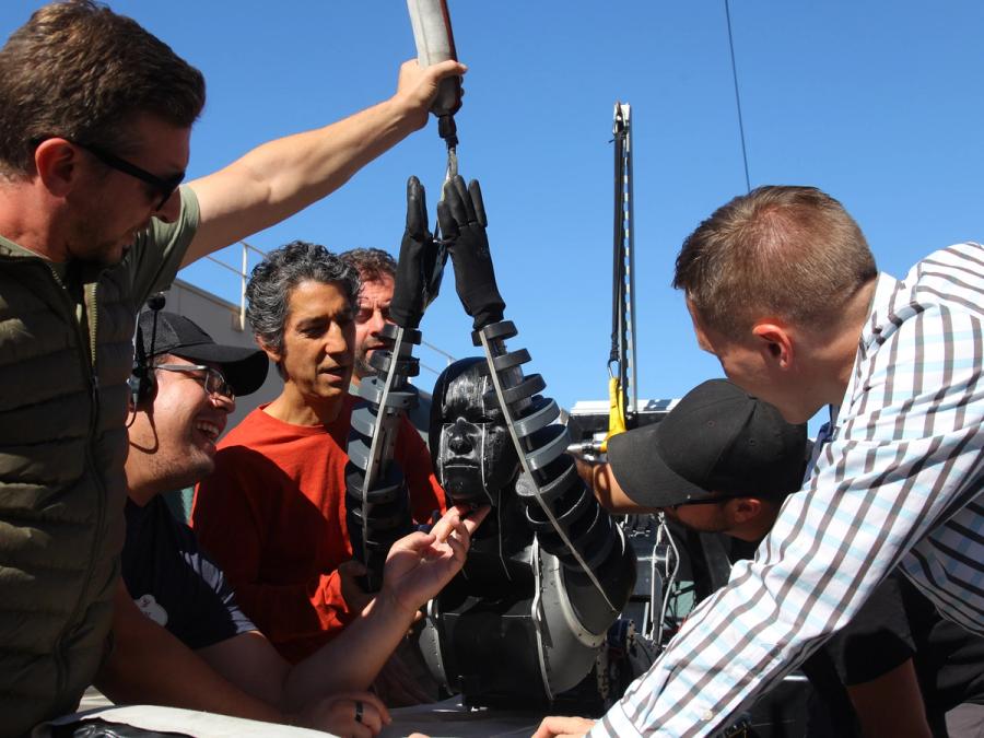 A group of people gather around a humanoid robot that is on it's stomach with its torso and head being lifted by a tether attached to its arms.