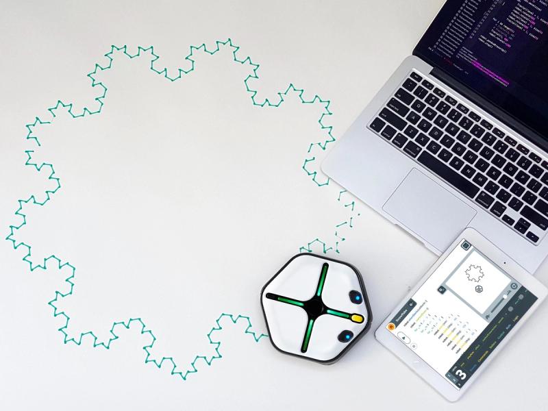 A squat white hexagonal robot with glowing lights sits in front of a tablet and laptop. It has drawn a large shape like a snowflake.