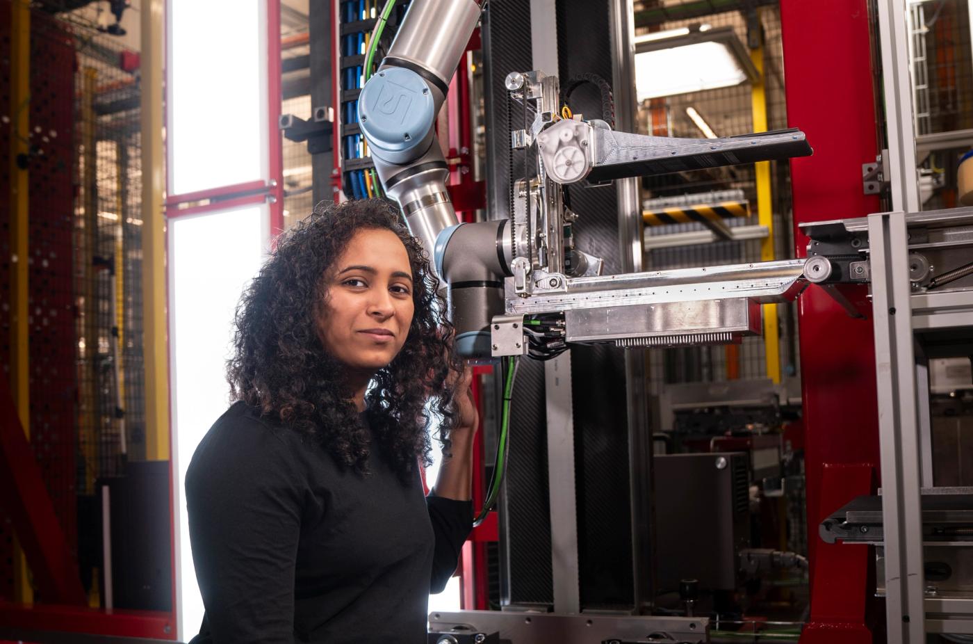 A woman with dark curly hair and a black shirt kneels in front of a robotic system at Amazon.