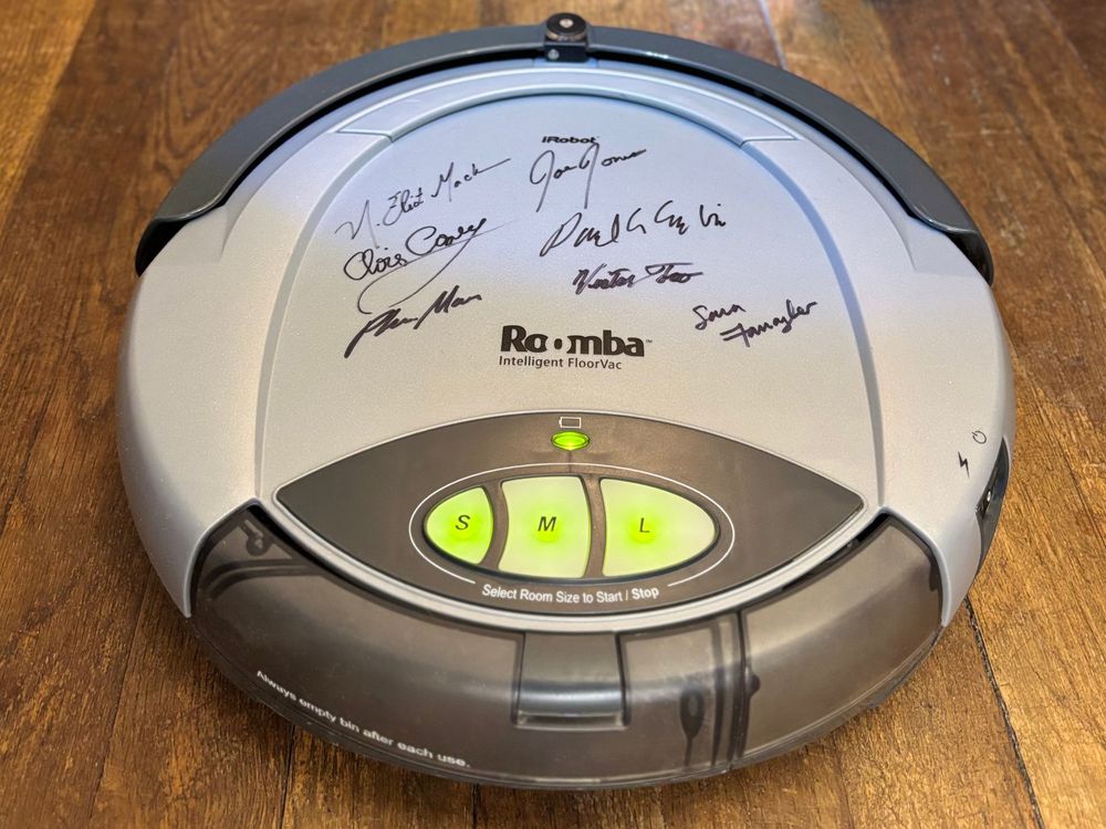 Roomba vacuum signed by iRobot team, showing control buttons, on wooden floor.