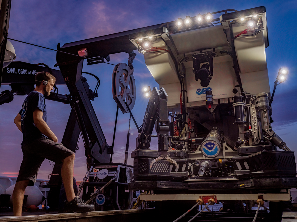 a person stands on a boat in front of a large mechanical apparatus bearing NOAA logo
